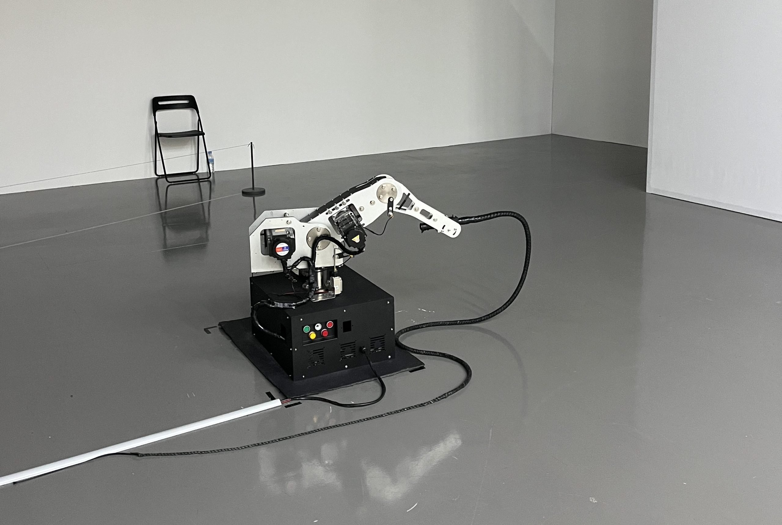 A robot-like arm holds a black whip in a large room with white walls.