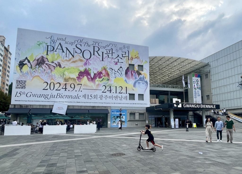 A boy on a two-wheel scooter roaming in a plaza with grey tiles in front of a big building with a banner that reads Gwangju Biennale.