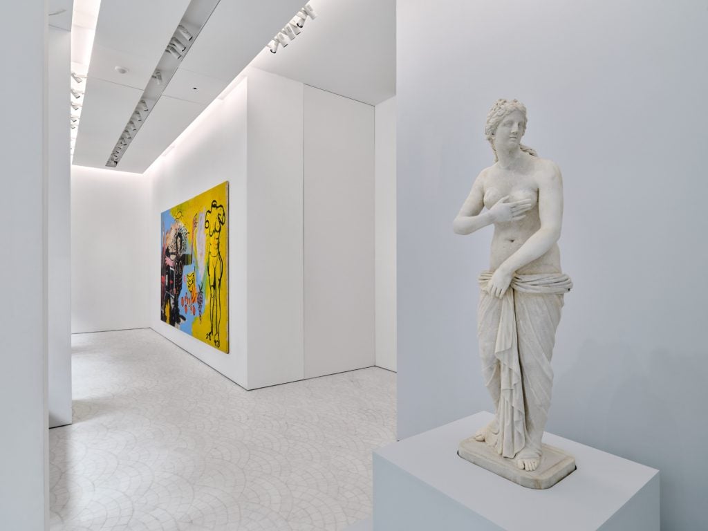 an interior of a brightly lit white-walled gallery space in which in the foreground is installed an ancient Roman marble statue of a female figure draped in a cloth and clutching her breast, hanging on the wall in the background seen from a side angle is a painting in shades of black, blue, and yellow