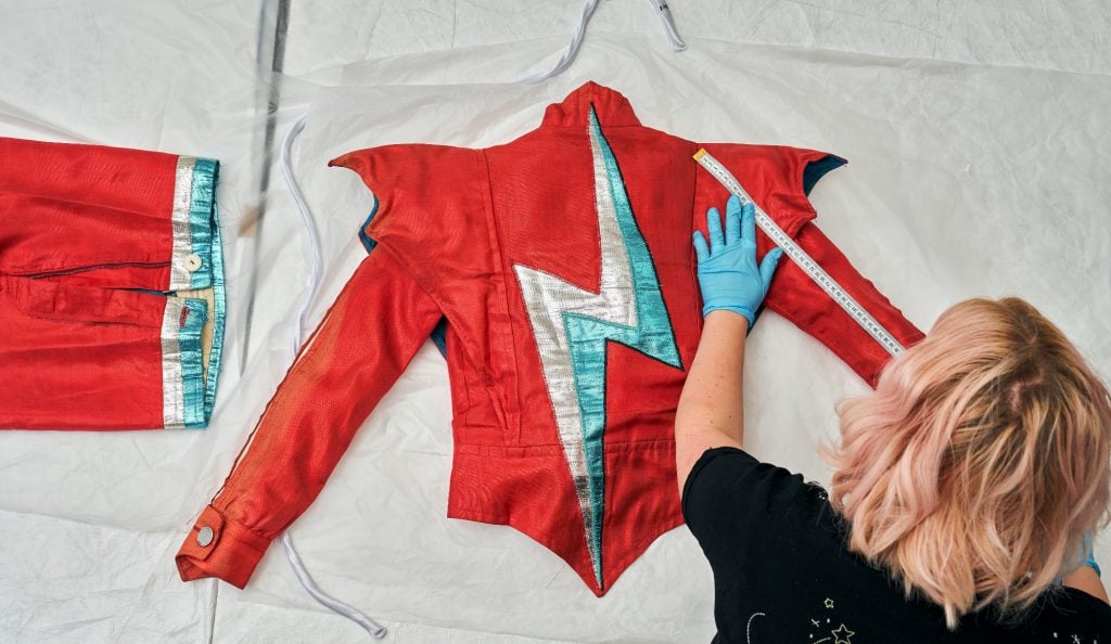 a woman seen from above leaning over a table with blue gloves touching a red jacket with a silver and blue stroke of lighting running down the back