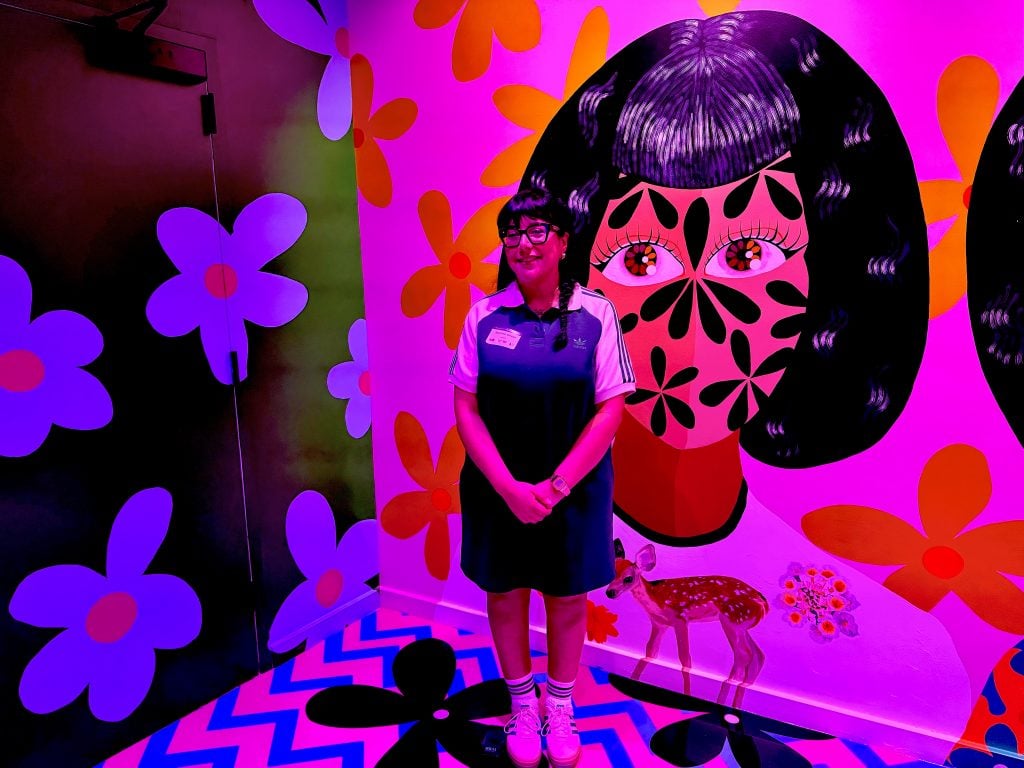 Jasmine Zelaya with her art installation at Meow Wolf Radio Tave. She is a young brown women standing in front of a colorful cartoon mural of a woman's face, covered with black flower petals. 