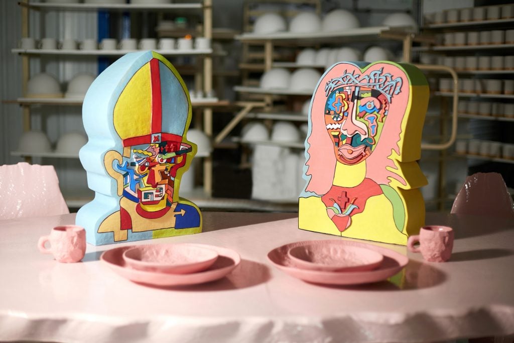 Two psychedelically decorated busts of Jesus and the Pope sitting at a table laid out with pink ceramicware