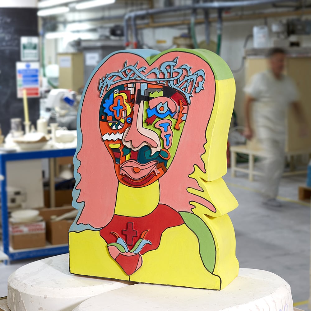 A colorful bust of Jesus by Robbie Williams, the subject's face filled with psychedelic panels, sitting on a stool in a workshop