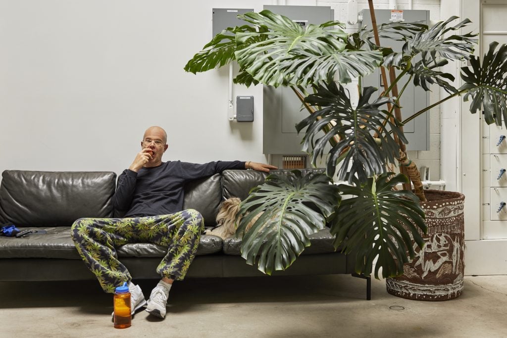 a white man in a blue top and camo pants sits on a sofa about to take a bite of a piece of fruit. a large fern reminiscent of those that appear in his paintings, sits next to him on the floor