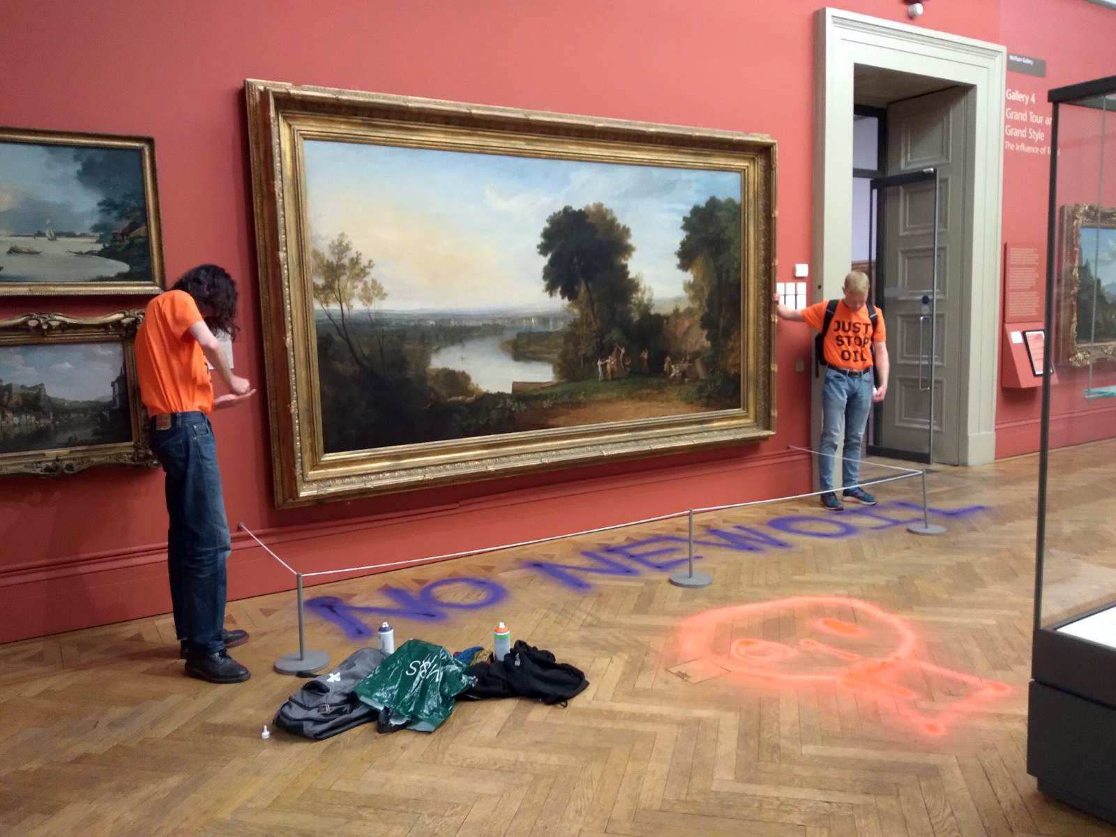 two young men in orange t-shirts stand by a large traditional landscape painting in a museum, one has his hand attached to the frame and the other is putting something on his hand. "no new oil' is spray painted in purple on the floor below the painting.