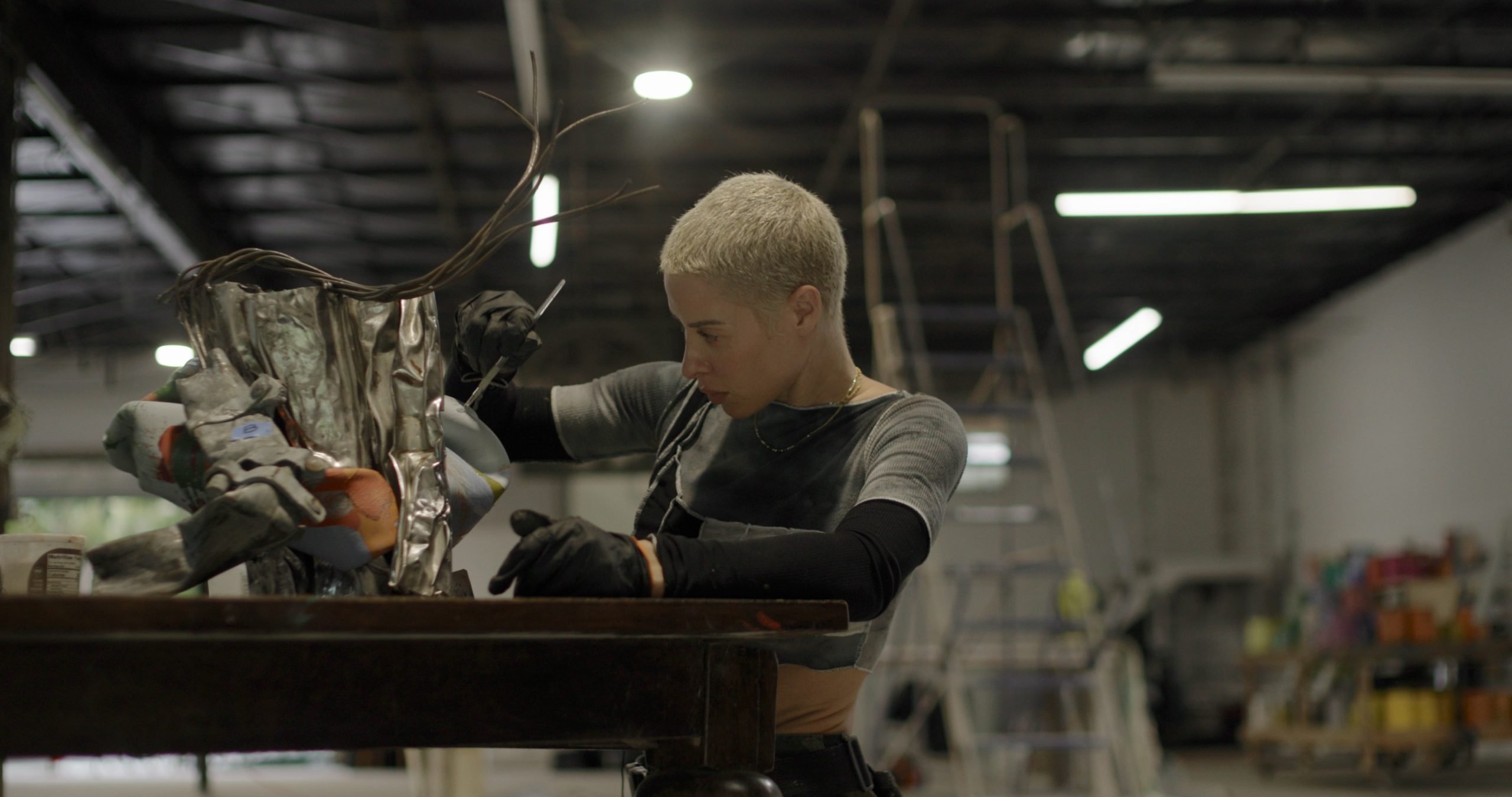 a woman with short blond hair works in an industrial studio on a metal sculpture