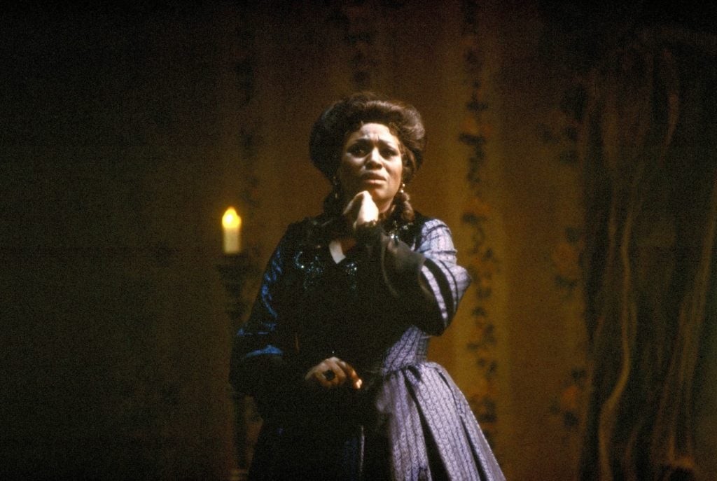 A woman standing in a dimly lit room, dressed in a formal, dark-colored, period costume with long sleeves, adorned with delicate patterns and lace. Her expression is intense and dramatic