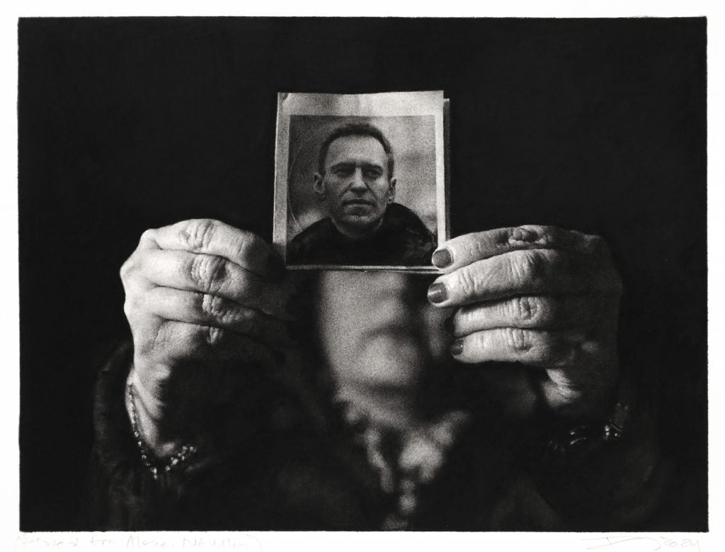 A black-and-white drawing showing two hands holding up a photograph of Russian opposition leader Alexei Navalny. The hands are detailed with deep shadows and textures, with the focus on the face of Navalny, framed by the hands.