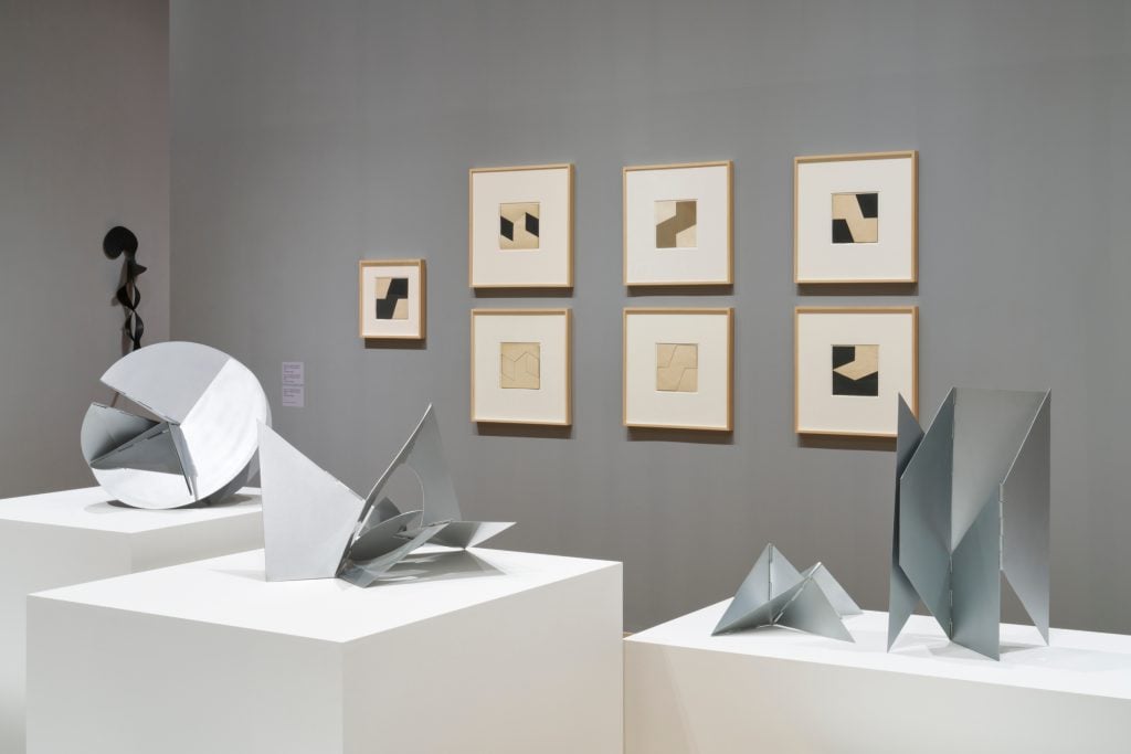 artworks installed in a gallery space with grey walls, in the foreground are abstract sculptural forms on white podiums, in the background are seven small framed geometrically abstract compositions hanging in a tiled manner on the wall