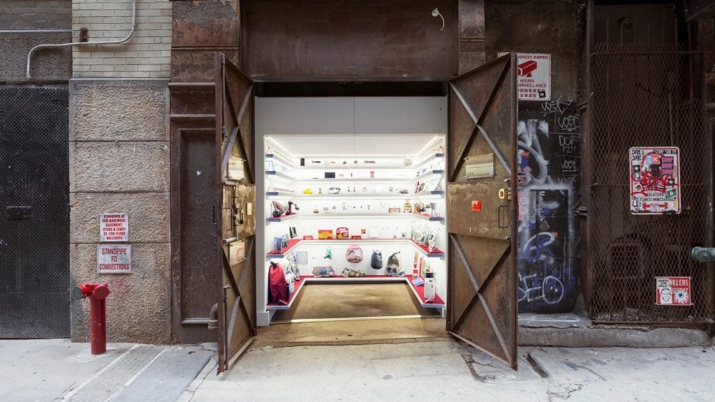 The Mmuseumm, a tiny white room lined with shelves displaying objects that opens directly to the street, with two double doors.