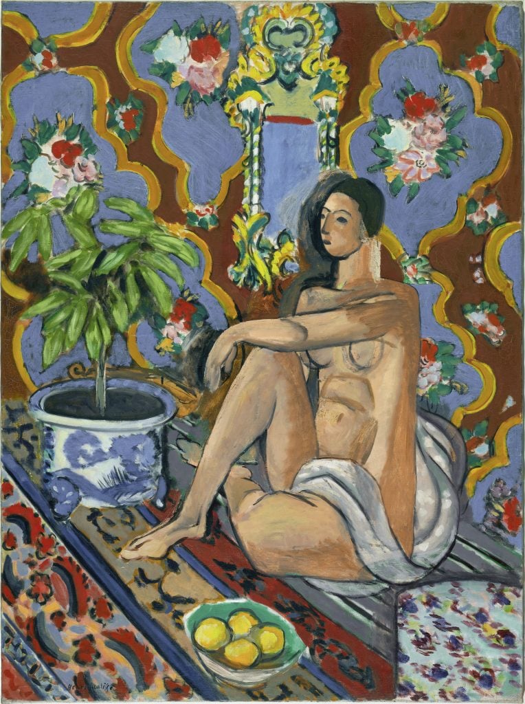 A woman in nude sitting on the floor with her legs crossed. She has dark hair, sitting against a colourful backdrop, with a bowl of yellow fruit in front of her and a green plant next to her