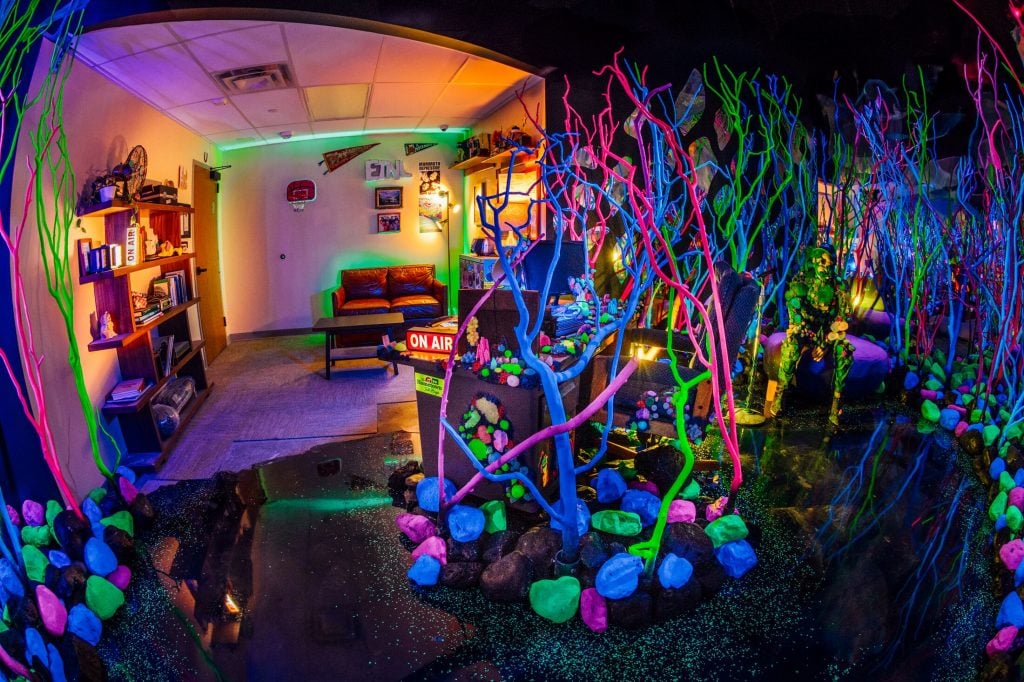 An office at Radio ETNL merges with the Gloquarium at Radio ETNL at Meow Wolf Radio Tave in Houston. The wide-angle lens photo should neon colored colar sculptures in a room illuminated by black light, with a nondescript radio office in the background.