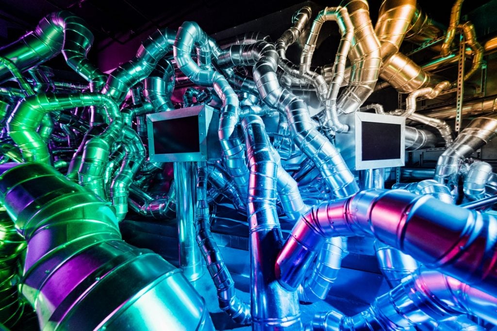 An art installation by Havel Ruck Projects at Meow Wolf Radio Tave in Houston. It is a tangle of silver AC vents connecting to two darkened monitors, the room lit by green, blue, and purple lights against a black background.