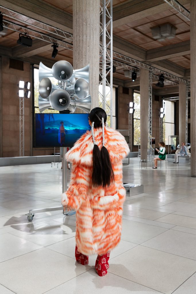 A Miu Miu model in a furry white and orange coat, with speakers behind her