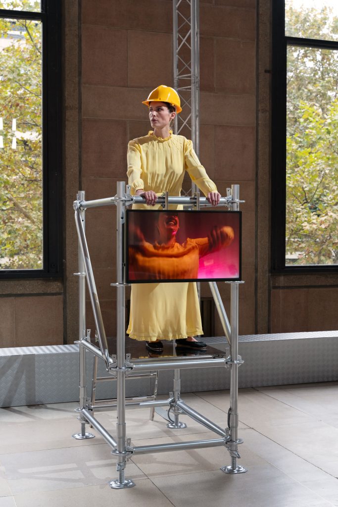 a woman in yellow in a hard hat on a scaffold featuring a tv screen