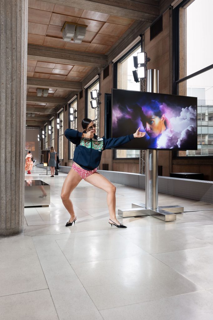 a woman in a black jumper and pink pants stands next to a tv screen