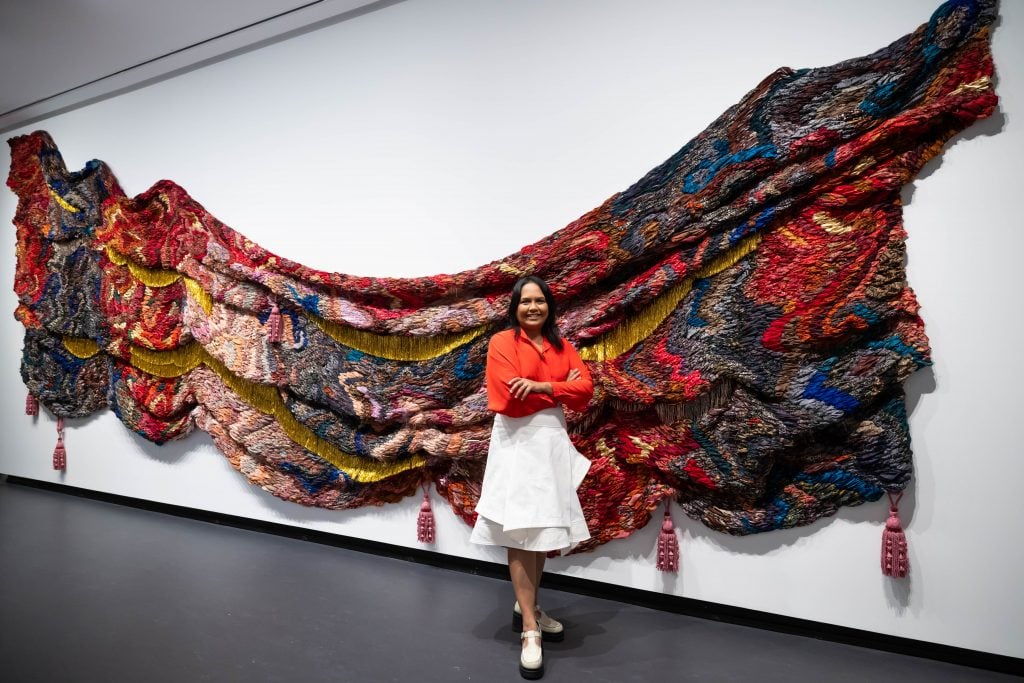 Suchitra Mattai with her piece a cosmic awakening (2023), at her show “Suchitra Mattai: Myth From Matter” at the National Museum of Women in the Arts, Washington, D.C. A large fabric tapestry made of multicolored saris and gold tinsel mounted on a white wall. Five beaded tassels hang from the bottom of the fabric. The artist, an Indian woman with dark hair wearing a red long-sleeved collared shirt and a large white skirt to below the knee with white leather platform heeled shoes, stands in front of the artwork.