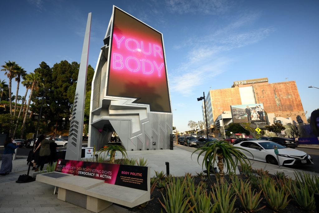 Nancy Baker Cahill, Body Politic (2024) on a billboard on the Sunset Strip in Hollywood. A vertically oriented billboard shows an image of a pink neon sign reading 