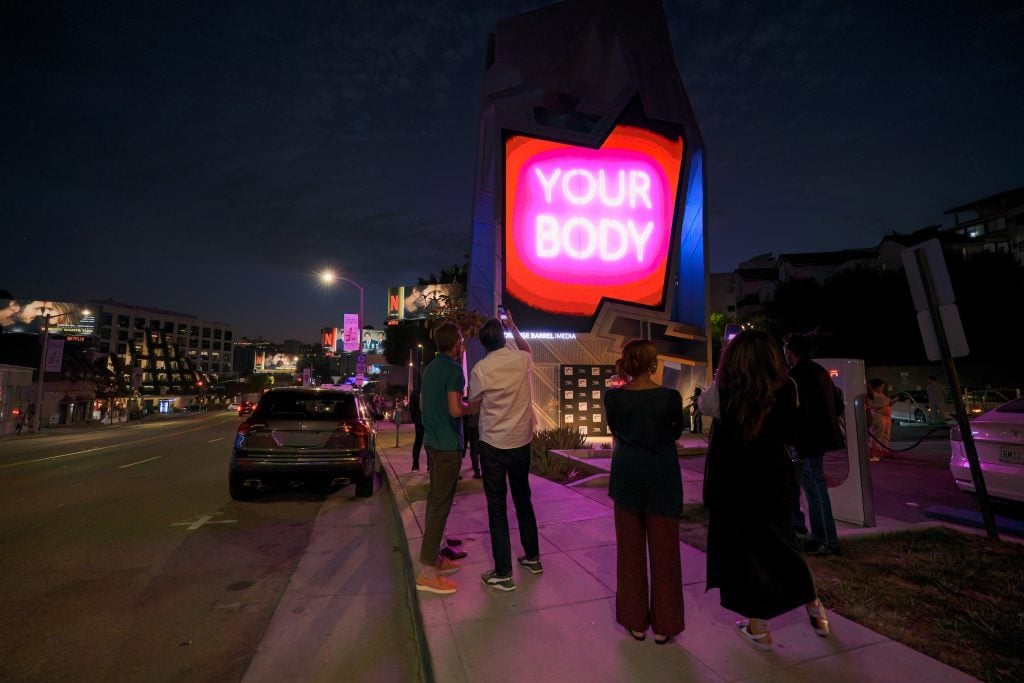 Nancy Baker Cahill, Body Politic (2024) on a billboard on the Sunset Strip in Hollywood. A billboard shows an image of a pink neon sign reading "Your Body" against a dark night sky.