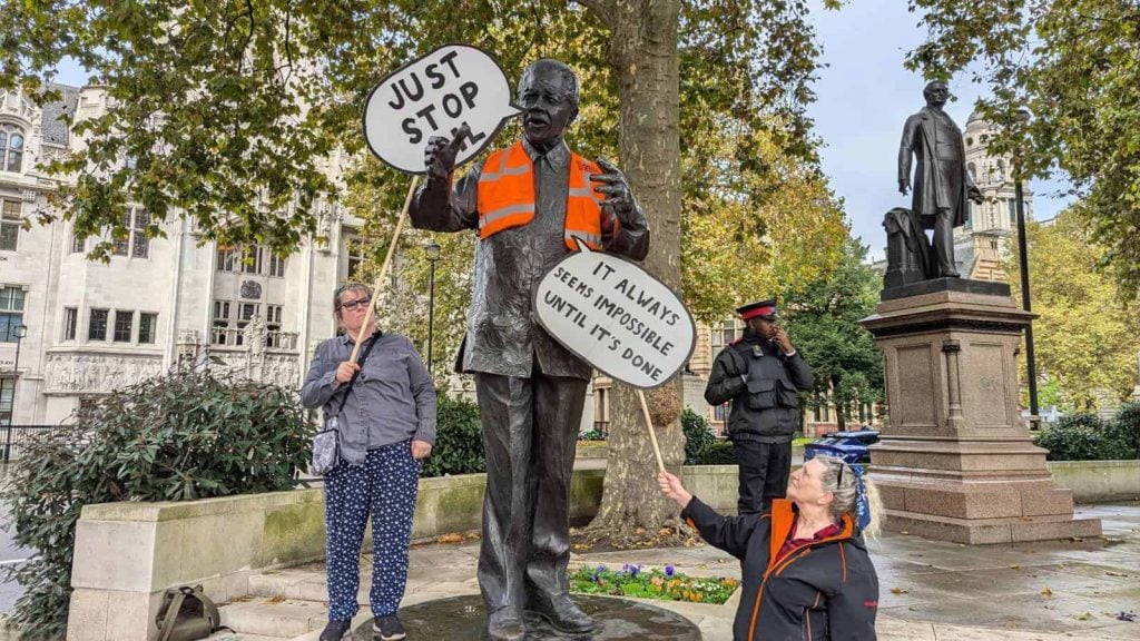 outside in a public spare with threes is a bronze statue of a man who has been draped in an orange high-vis jacket and next to him two women hold up speech bubble-shaped signs reading 'just stop oil' and 'it always seems impossible until it's done'