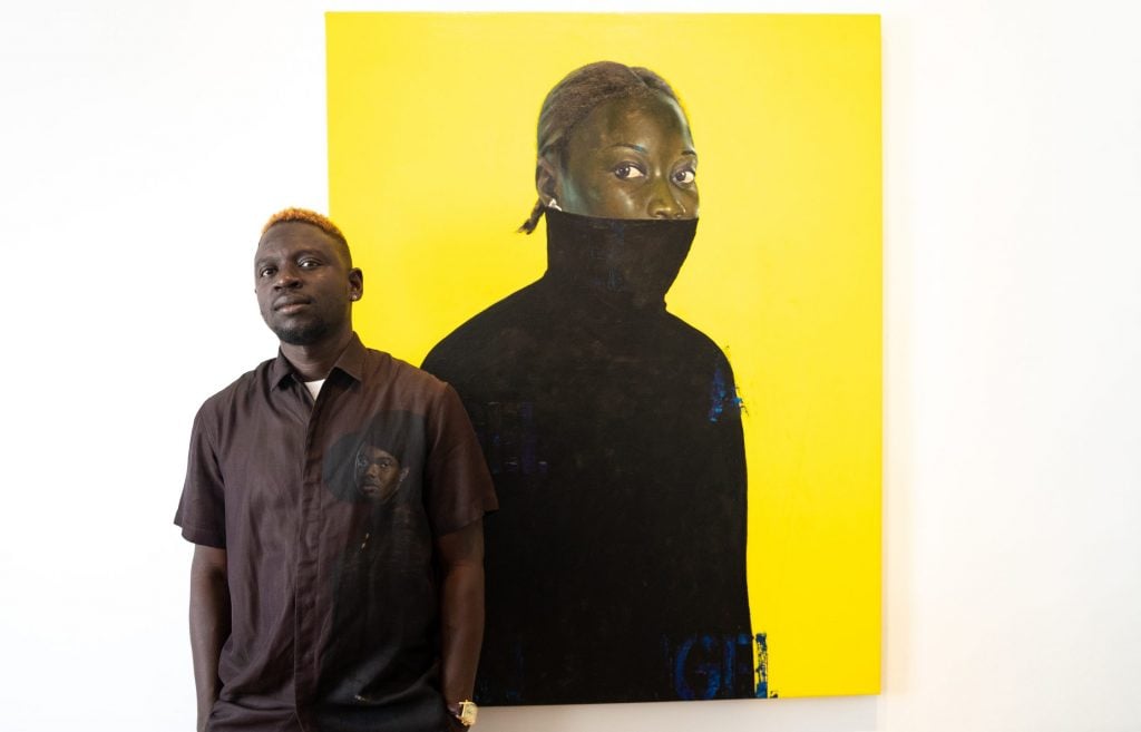 A man of African descent posing next to a painting of a Black person with half of their face covered, against a yellow backdrop.