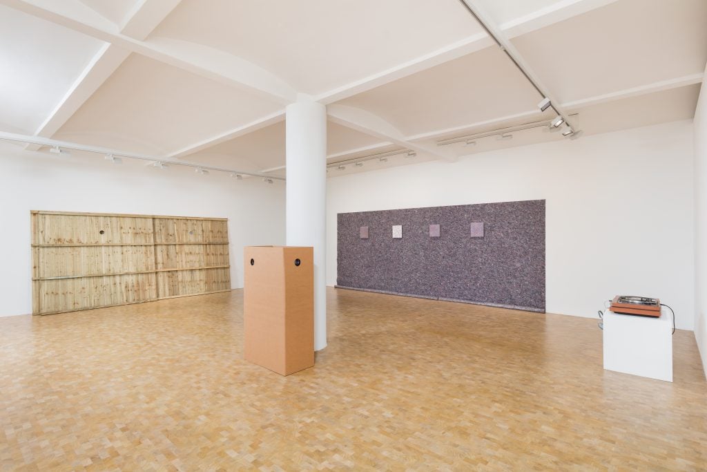 Art gallery with wooden panel installation, cardboard sculpture, textile wall art, and turntable setup