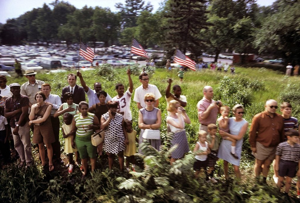 Paul Fusco, RFK Funeral Train (1968). A color photo of crowds watching the train go by, taken from the train and slightly blurry from the motion of the car. Three people in the center hold up American flags. 