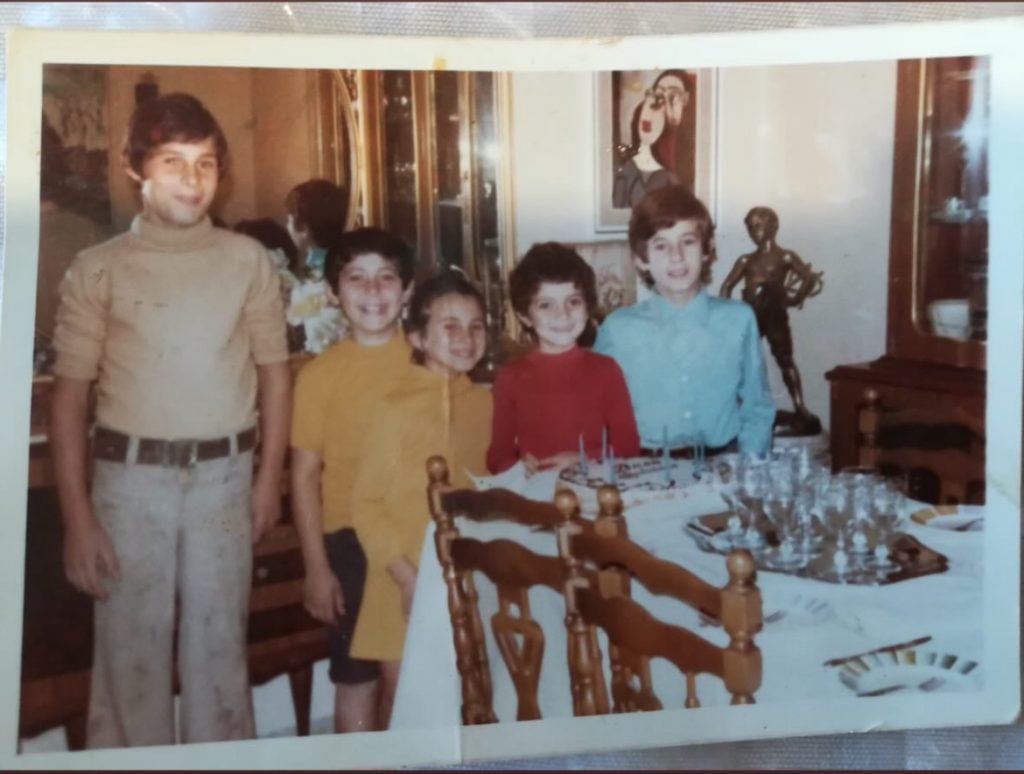 five smiling children stand in a family home with a dining table in front of them, behind there is a painting of a woman on the wall, the photograph was clearly taken many decades ago