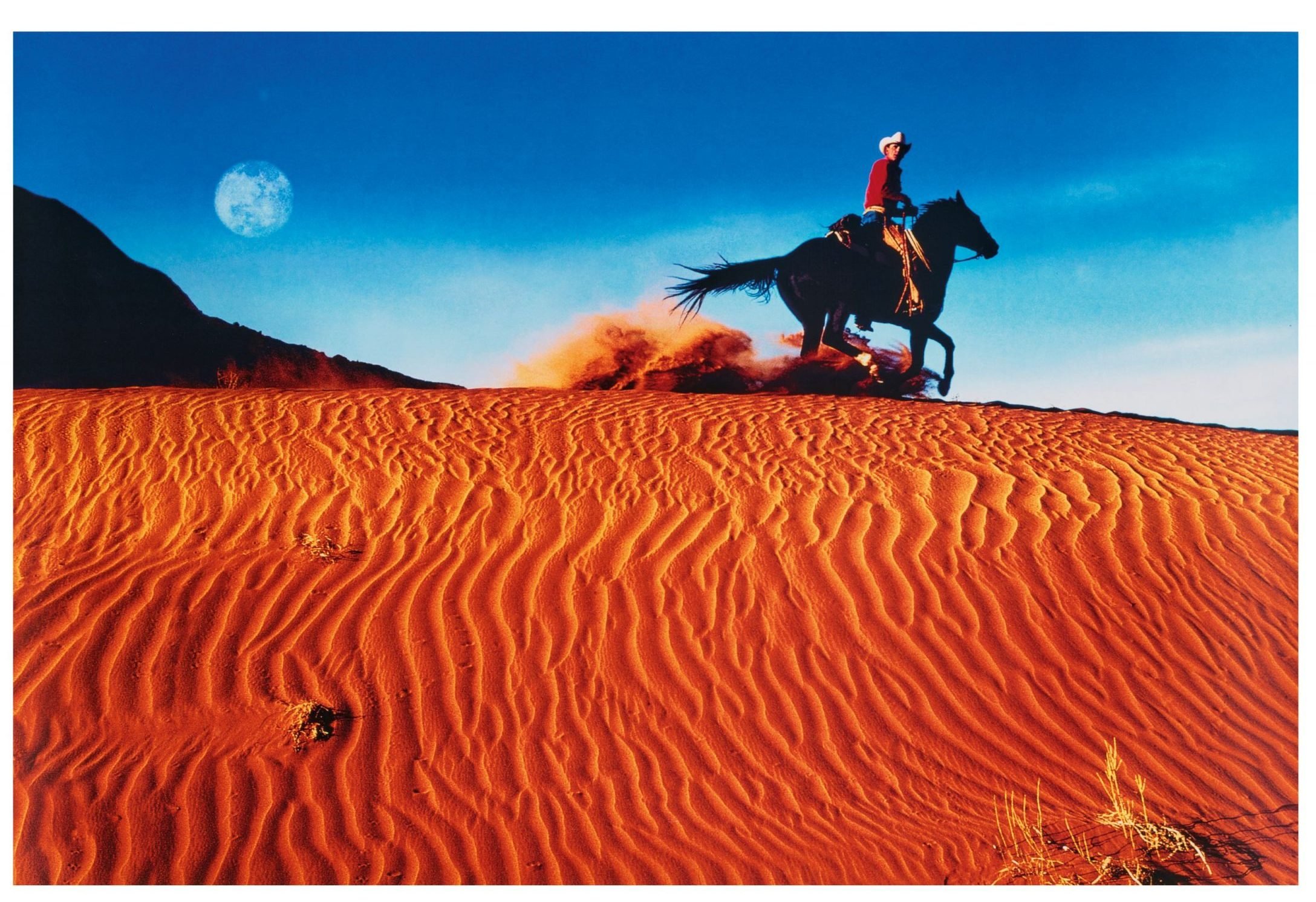 A color photo shows a cowboy on a horse kicking up orange sand