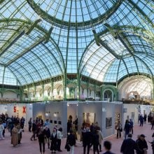 A large crowd explores an art exhibition inside a grand, glass-domed hall with intricate green ironwork and natural lighting
