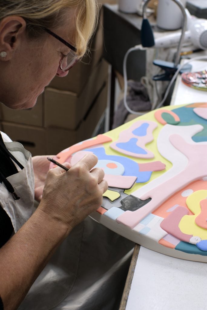 A woman sitting at a table painting a large ceramic slab in bright colors