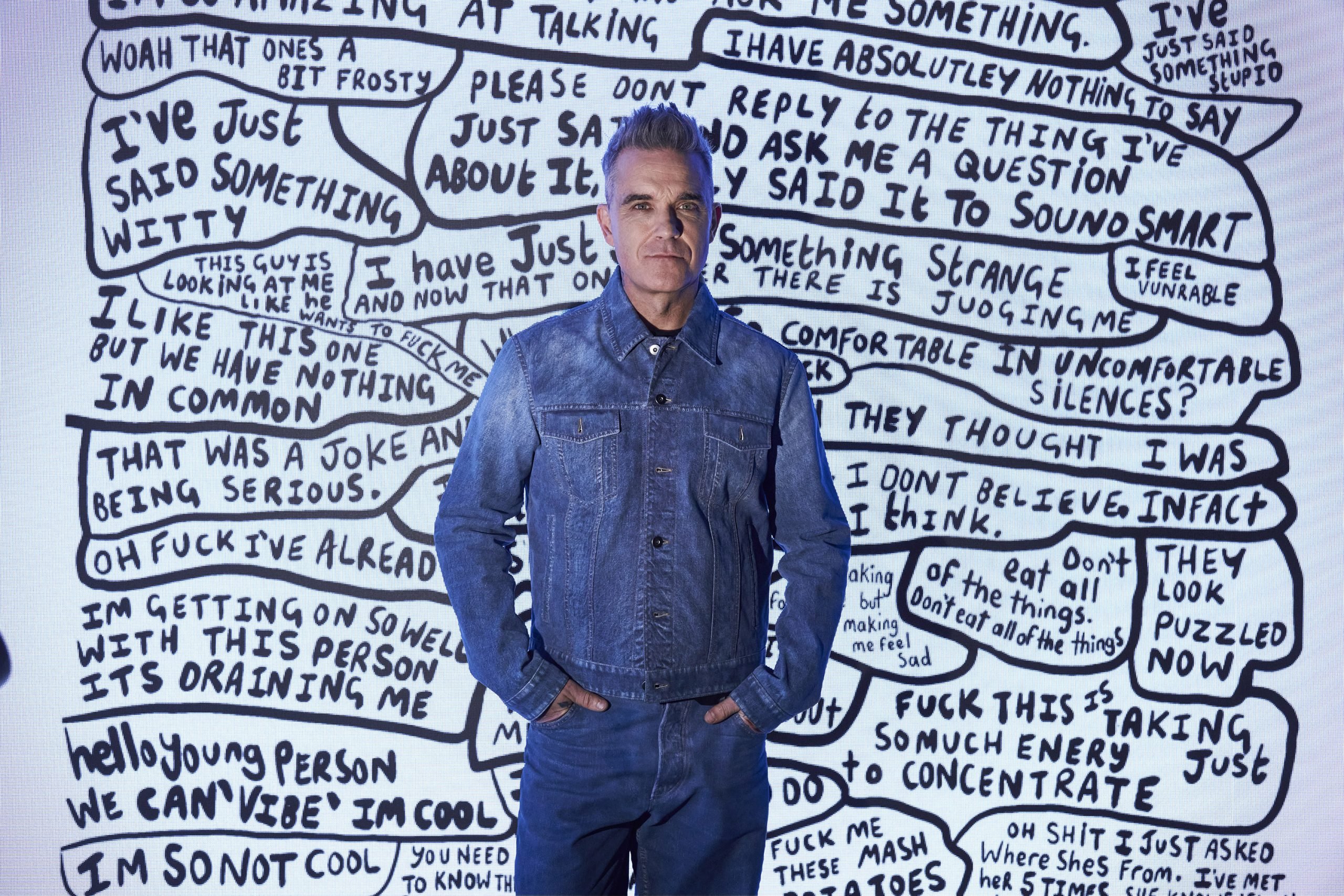 A man in a denim jacket and jeans standing in front of a screen showing handwritten slogans in bubbles