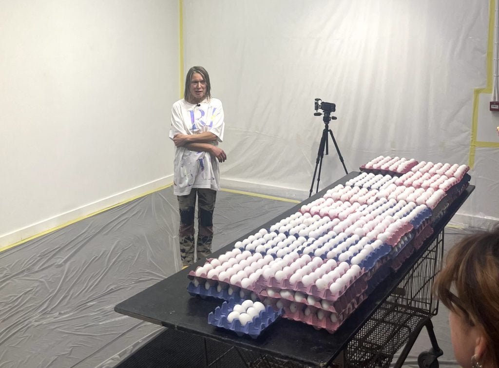 a woman in a large white t-shirt stands in a white-walled room next to a table covered in layered trays filled with white eggs