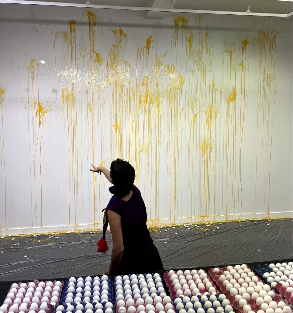 a woman hurls an egg at a white wall stained with long drips of egg yolk from previously thrown eggs. There are crushed egg shells on the floor beneath. in the foreground there are many trays of white eggs