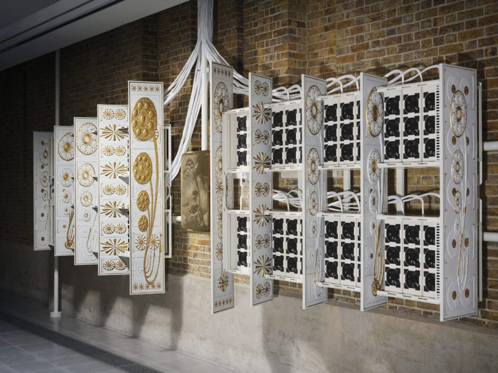 a large organ-lke structure that is white with gold detailing and has black GPU's installed in the gaps is mounted onto the brick wall of a gallery space