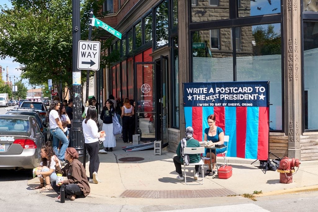 Sheryl Oring, I Wish to Say at Co-Prosperity in Chicago during the Democratic National Convention in August 2024. A woman dressed like a 1950s secretary sits at a makeshift desk on the street corner in front of a typewriter and a red and blue striped banner inviting the public to send a letter to the next president. 