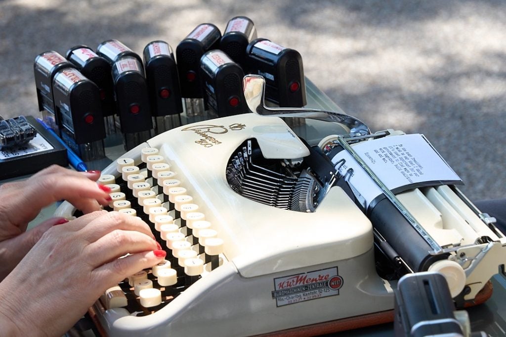 Sheryl Oring, I Wish to Say. A close up shot of the artist's hands, with red nail polish, typing on a vintage white typewriter. 