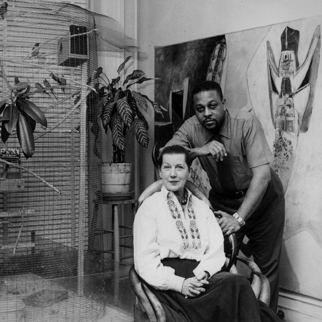 a black and white photo from the 1950s shows a man and woman in an apartment next to a large birdcage, they are the artist couple Jeanne Reynal and Thomas Sills 