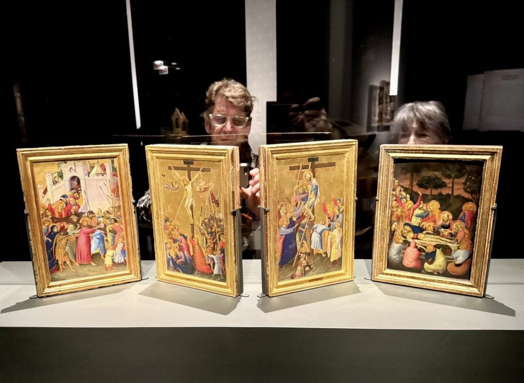 Two people are reflected in glass while viewing four medieval religious paintings of Christ’s Passion