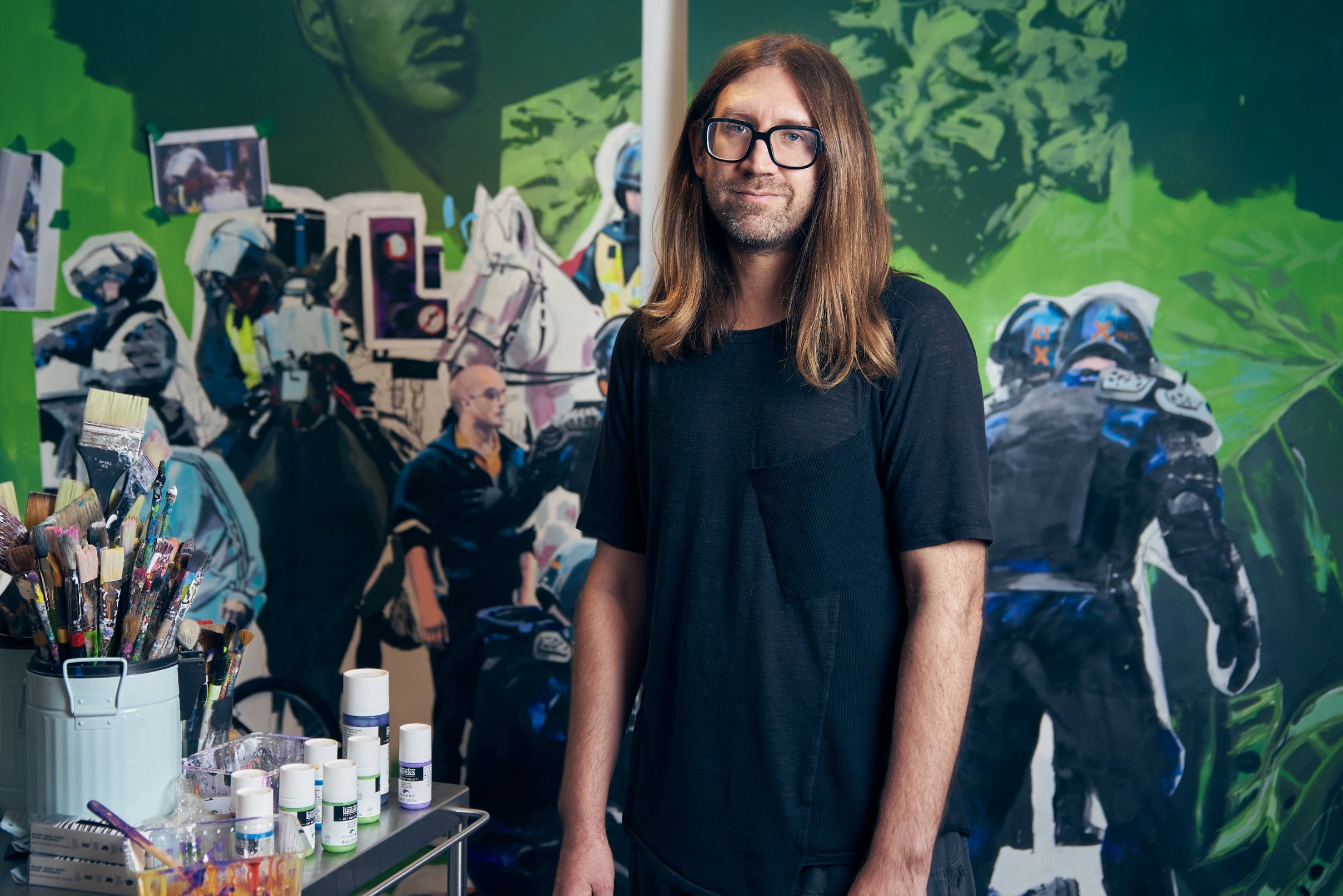 A portrait of the artist Stuart Semple in his studio. He is a man in his early 40s, with long straight light brown hair, wearing thick rimmed glasses and a black t-shirt. He is standing with his arms at his side, with a blank expression on his face. A cart of paintbrushes and paint is to his left and there is a green painting behind him featuring figures of police officers.