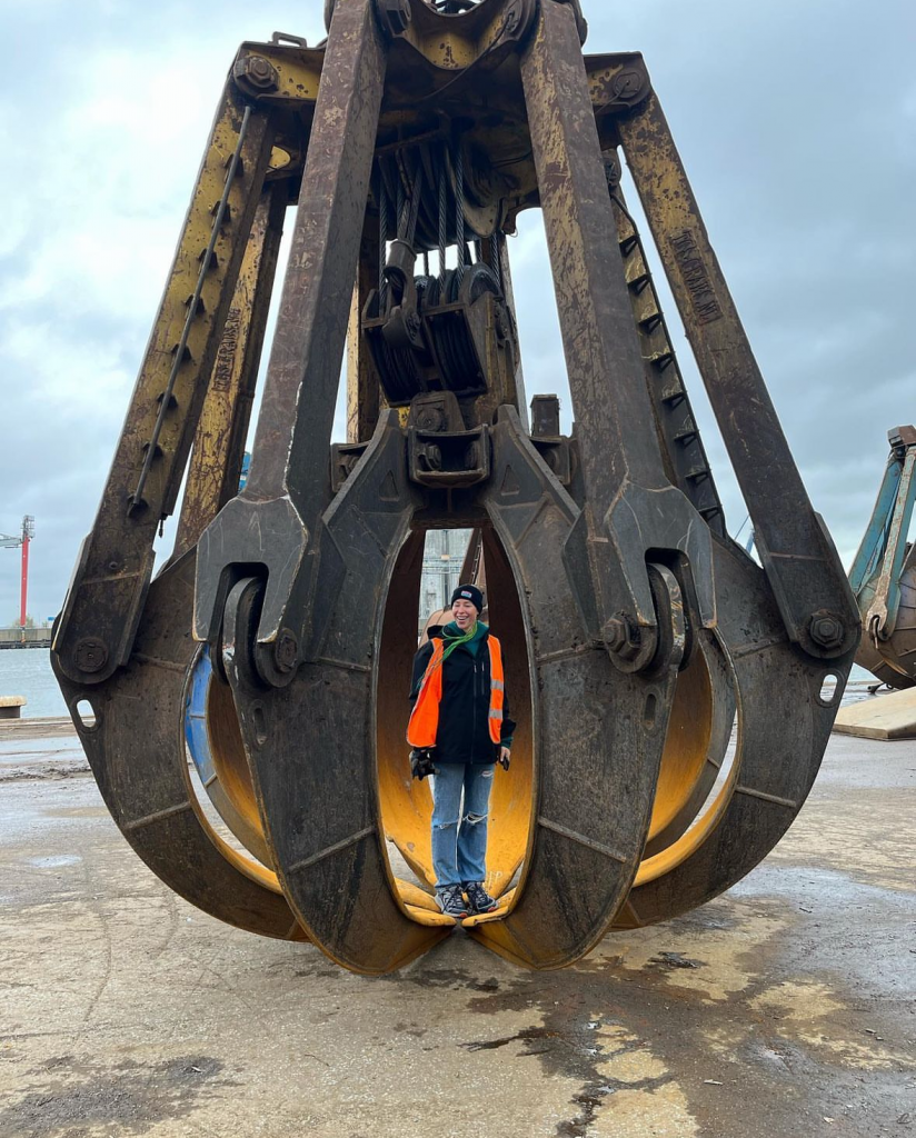 a woman in a high vis vest and jeans stands in the claw of a giant construction truck