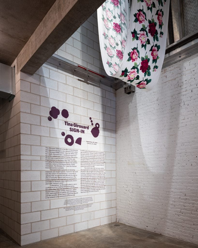 Fabric from Solomon's Lot hangs in "Tina Girouard: Sign In" at Center for Art Research and Alliances, New York. A long length of white fabric with a large print of pink and red roses hangs from in ceiling in the top of the photograph. Below, there is introductory wall text for the show.