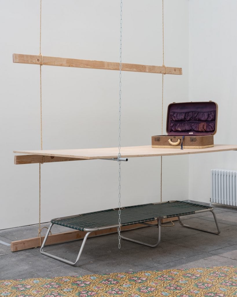 Tina Girouard, Hung House (1971) on view "Tina Girouard: Sign In" at Center for Art Research and Alliances, New York. A cot lies on the floor, beneath a suspended plywood panel on top of which sits and open suitcase.