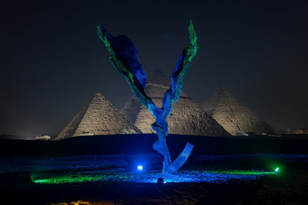 a winged creature lit in blue and green is situated in front of the pyramids