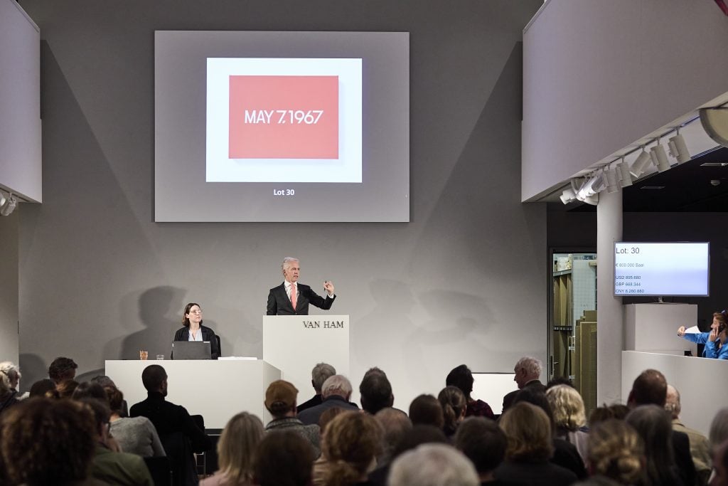 an auctioneer stands at a podium and auctions a large red painting