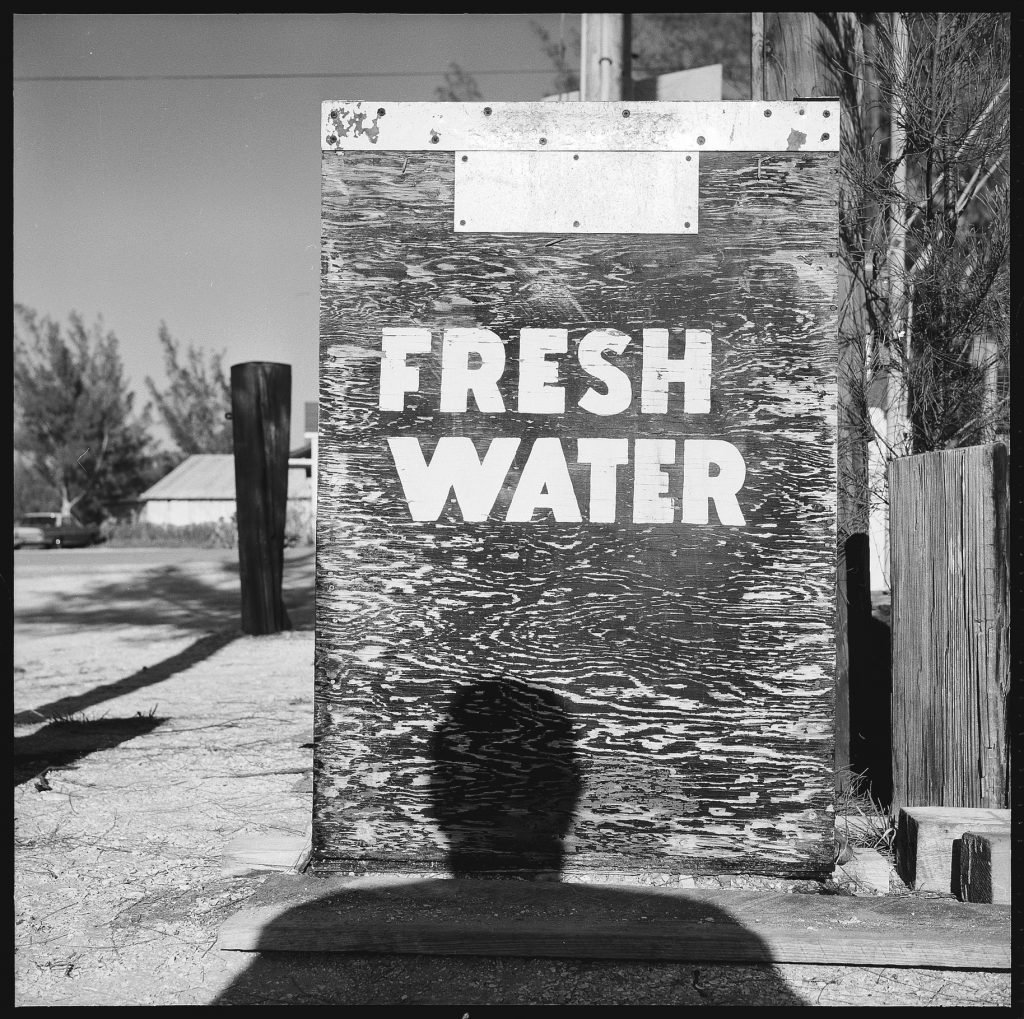 A weathered sign reading "FRESH WATER" with a shadow of a person cast on it.