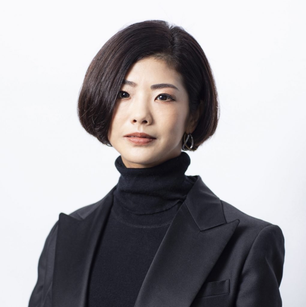 An East Asian woman in short hair and black outfit, against a white background