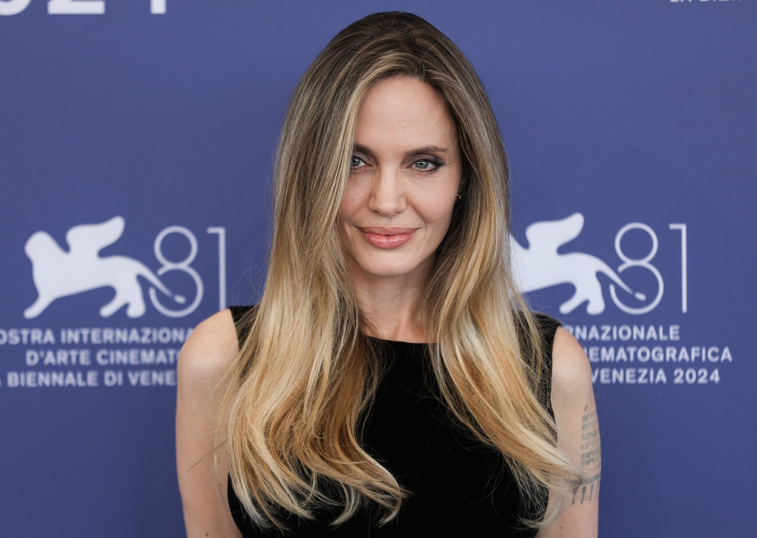 Actor Angelina Jolie, pictured with dirty blonde hair and in a sleeveless black top, posing against a blue backdrop