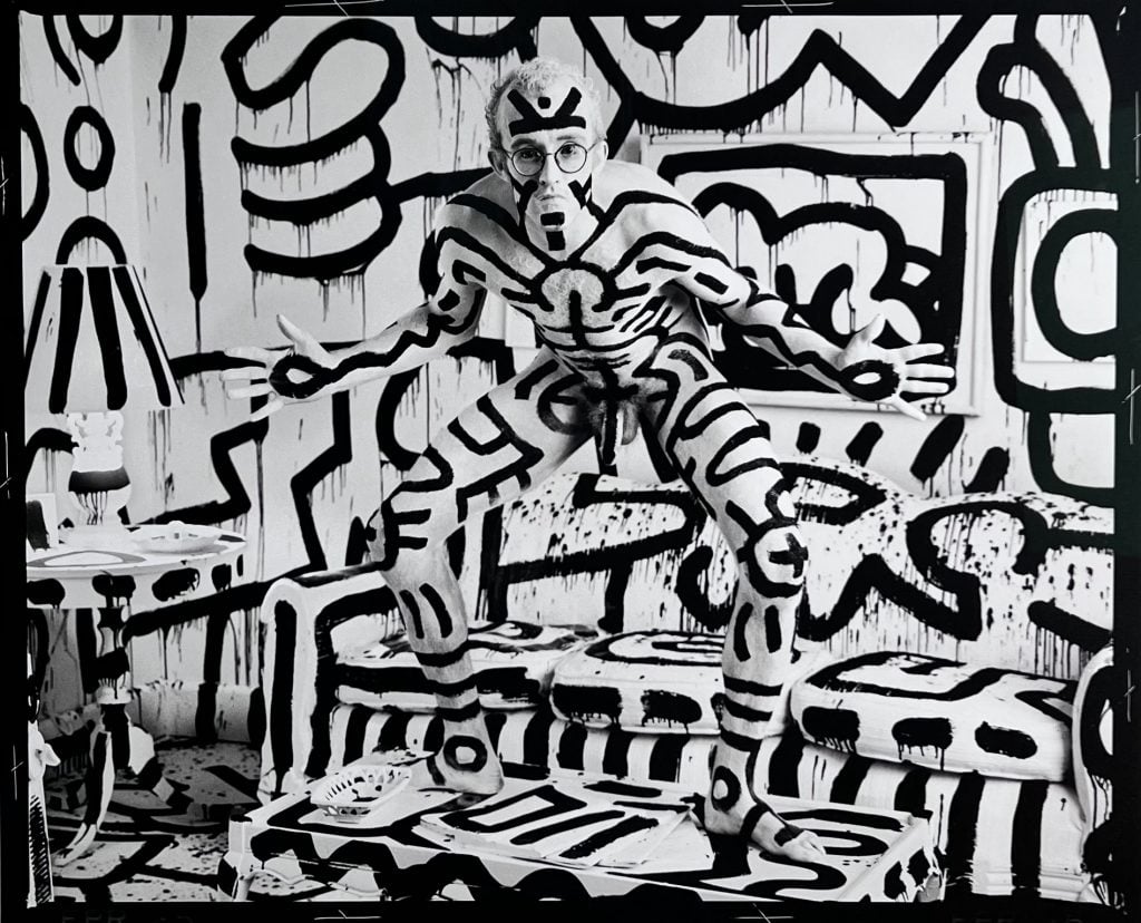 Black and white photo of artist Keith Haring nude standing on a coffee table in a living room where the entire room, objects, furniture, as well as himself are painted in his signature doodle pattern, included in the Important Photographs sale.