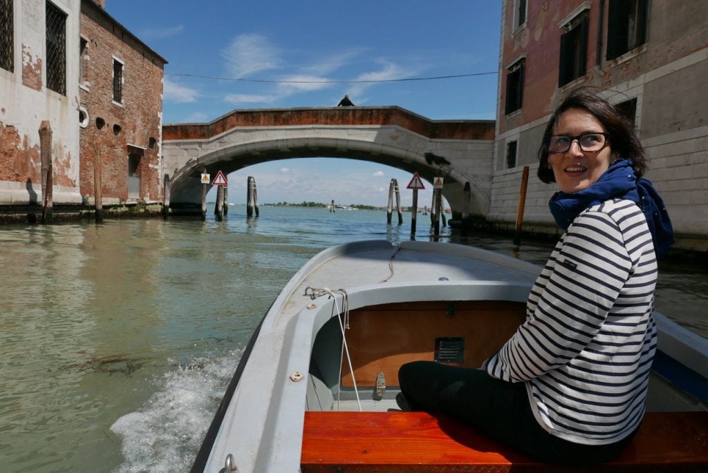 Artist Melissa McGill wearing a stripped shirt sitting in the front of a gondola as it approaches one of the city's bridges. The waterway is lined by old-world brick buildings.